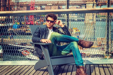 Adam New York'ta seyahat. Giyinme siyah deri ceket, kot, kahverengi önyükleme ayakkabıların, güneş gözlüğü, sakallı, güverte, sandalyede oturan genç bir adam rahatlatıcı. Tekneler, arka plan köprüde