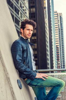 Serious European businessman in New York. Wearing black leather jacket, blue jeans, a young guy with beard, leaning against wall, facing surrounded high buildings, frowned, unhappy, thinking clipart