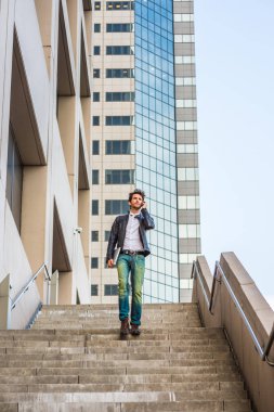 Merdivenlerden inip çalıştıktan sonra ofis dışında eve yürürken cep telefonundan ailesine söz giyen deri ceket, kot pantolon, dizüstü bilgisayar, taşıyan kol genç profesyonel.