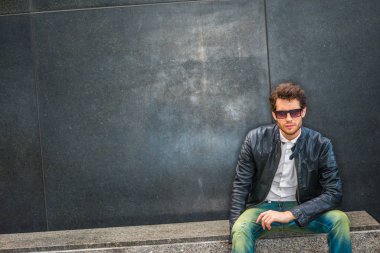 Siyah deri ceketli açılarak, beyaz gömlek, kot, altında soyunma giymiş güneş gözlüğü, karşı mermer bankta oturmuş sakallı genç adam köşedeki sokak, üzgün, depresif, düşünme üzerine duvar