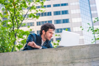 New York'ta eğitim Avrupa yüksek lisans öğrencisi. Siyah deri ceketli, sakallı, okuma, dizüstü bilgisayar duvar üstünde iş bölgesi üzerinde bahar günü içinde çalışan genç bir adam
