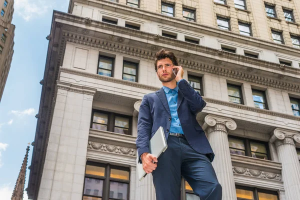 Power of Technology in our modern daily life. Dressing in blue suit, a young European Businessman with beard standing outside in New York, arm carrying laptop computer, talking on mobile phone
