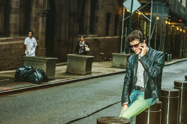 Siyah deri ceket, kot, kahverengi çizme ayakkabı, güneş gözlüğü, New York, sokakta oturan sakallı genç bir adam takıyor gülümseyen, telefonda konuşurken, rahatlatıcı.