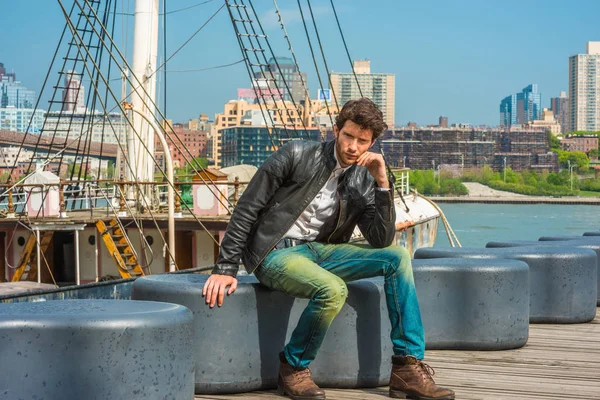 Man Spring Casual Urban Fashion. Wearing black leather jacket, blue jeans, brown leather boot shoes, a young guy with beard, sitting on bench at harbor, relaxing, thinking. A boat on background