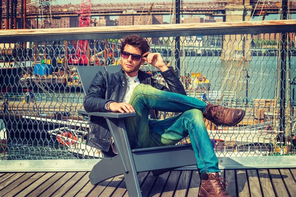Adam New York'ta seyahat. Giyinme siyah deri ceket, kot, kahverengi önyükleme ayakkabıların, güneş gözlüğü, sakallı, güverte, sandalyede oturan genç bir adam rahatlatıcı. Tekneler, arka plan köprüde