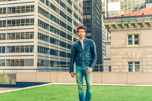 European graduate student studying in New York. Wearing black leather jacket, blue jeans, carrying laptop computer, young guy with beard, standing on lawn in business district, looking forward