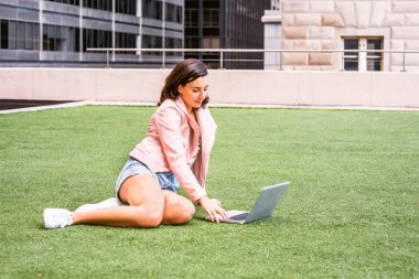 Genç Amerikan Koleji öğrenci eğitim n üzerinde hafif pembe deri ceket, NewYork, kot şort, beyaz spor ayakkabı, bacaklar, kampüste, yeşil çim üzerinde oturan dizüstü bilgisayarda çalışarak bükme sökük