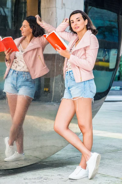 若いアメリカ人女性留学ニューヨークで勉強して 身に着けている光ピンクのレザー ジャケット 青の破れたデニムのショート パンツ 白いスニーカー キャンパスで反射と鏡のそばに立って読書赤ブーします — ストック写真