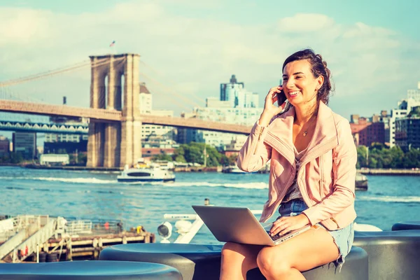 若いアメリカ人女性旅行 ニューヨークで仕事取り組んでラップトップ コンピューター 携帯電話で話す東川のそばに座って笑顔します マンハッタン橋 ブルックリンの建築物 遠くの背景にボート — ストック写真