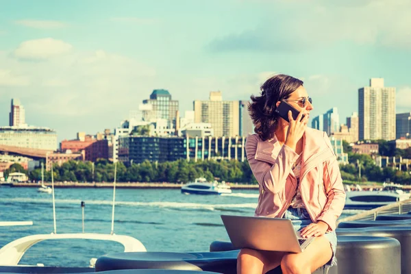 若いアメリカ人女性旅行 ニューヨークで仕事サングラス 川のそばに座って ラップトップ コンピューターで作業して 周りを探して 携帯電話で話しています ブルックリンの建物 背景のボート — ストック写真