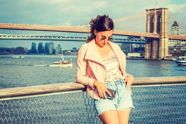 Young American Woman traveling in New York, wearing pink leather jacket, blue Denim shorts, sunglasses, standing by river, looking down, thinking. Brooklyn, Manhattan bridges, boats on background