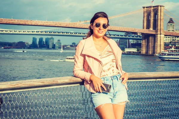 Young American Woman traveling in New York, wearing pink leather jacket, blue ripped Denim shorts, sunglasses, standing by river, looking, smiling. Brooklyn, Manhattan bridges, boats on background
