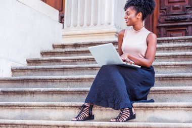 Genç Afrikalı-Amerikalı kadın New York'ta vintage ofis dışında merdivenlerde oturan kolsuz bluz, siyah etek, sandaletler giyen afro saç modeli ile dizüstü bilgisayar üzerinde çalışan, uzağa arıyorsunuz, gülümseyen
