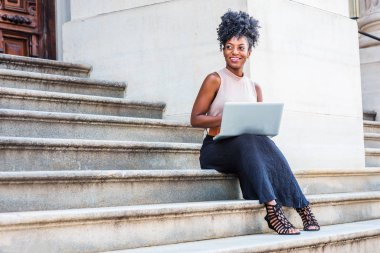 Başarı için yol. Dizüstü bilgisayarda çalışarak New York'ta ofis dışında merdivenlerde oturan kolsuz bluz, siyah etek, sandaletler giyen genç Afro-Amerikan kadın afro saçlı gülümseyen