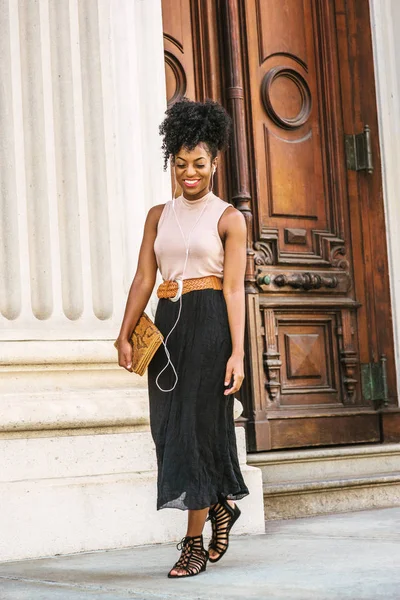 Young black woman with afro hairstyle wearing sleeveless light color top, black skirt, carrying small leather briefcase, listening music with earphones, walking by vintage office doorway in New York