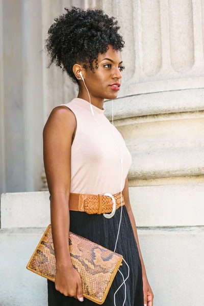 Young black woman with afro hairstyle, wearing sleeveless light color top, belt, carrying small leather briefcase, standing by column outside office building, listening music with earphones, thinking