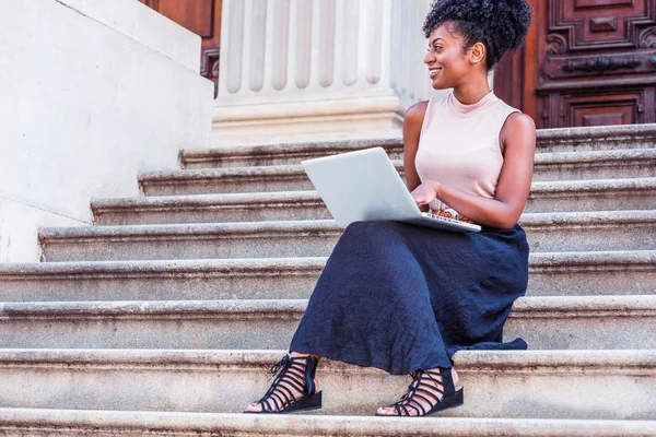 Genç Afrikalı-Amerikalı kadın New York'ta vintage ofis dışında merdivenlerde oturan kolsuz bluz, siyah etek, sandaletler giyen afro saç modeli ile dizüstü bilgisayar üzerinde çalışan, uzağa arıyorsunuz, gülümseyen