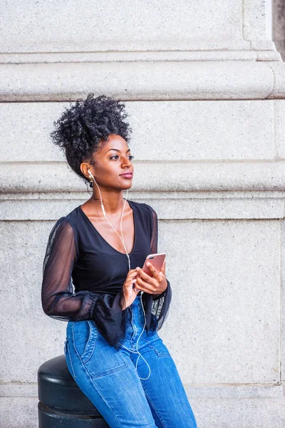 Young beautiful African American Woman with afro hairstyle wearing mesh sheer long sleeve shirt blouse, sitting on street in New York, listening music with earphone and cell phone, relaxing