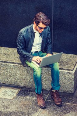 Siyah deri ceket, kot pantolon, çizme ayakkabı, güneş gözlüğü, Genç bir adam sakallı mermer bankta oturan giymiş, dizüstü bilgisayarda çalışarak Sigara İçilmeyen, çalıştıktan sonra sorunları çözmek için çaba sarf.