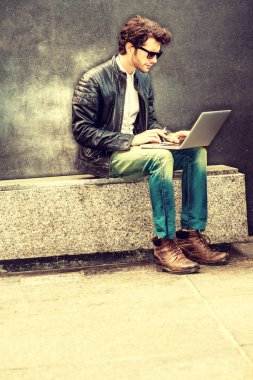 Deri ceket, kot pantolon, çizme ayakkabı, güneş gözlüğü, bir adam bankta oturan, Sigara içiyorum, dizüstü bilgisayarda çalışarak, çalıştıktan sonra sorunları çözmek için çaba sarf sakallı giyiyor. Retro filtre uygulanmış görünüm.