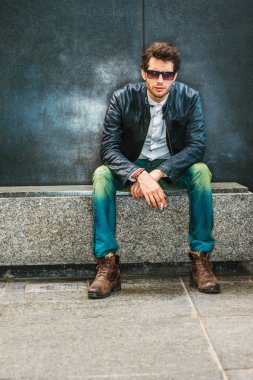 Giydiği siyah deri ceket, kot, kahverengi çizme ayakkabı, güneş gözlüğü, köşede, duman Sigara çalışma tatilinde, mermer bankta oturmuş sakallı genç bir adam sakin bir sakıncası çalışıyor