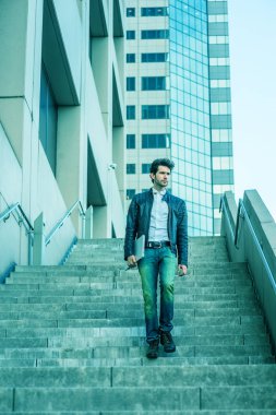 Deri ceket, kot pantolon, kol taşıma dizüstü bilgisayar, merdivenlerden inip office yoğun çalışma günün ardından yüksek bina dışında eve yürürken giyen genç adam sakallı. Mavi renk tonu ile filtre uygulanmış görünüm.