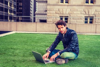 Seyahat, New York'ta çalışma Avrupa iş adamı. Siyah deri ceket, kot, önyükleme ayakkabılar giyen, sakallı, bir adam okuma, yeşil çim üzerinde otururken bacak bacak dizüstü bilgisayar üzerinde çalışan.