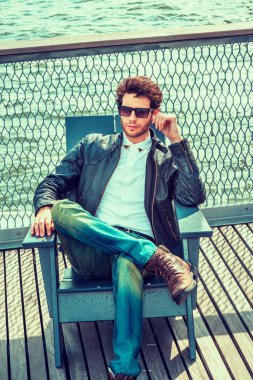 Avrupa New York'ta seyahat adam. Giyinme siyah deri ceket, beyaz gömlek, kot, kahverengi önyükleme ayakkabıların, güneş gözlüğü, güverte, sandalyede oturan sakallı bir adam bacak, bacak rahatlatıcı.