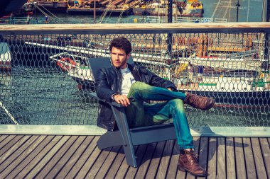 Avrupa New York'ta seyahat adam. Giyinme siyah deri ceket, kot, kahverengi çizme ayakkabı, sakallı, güverte, sandalyede oturan bir genç adam bacak, bacak rahatlatıcı. Tekneler arka plan üzerinde.