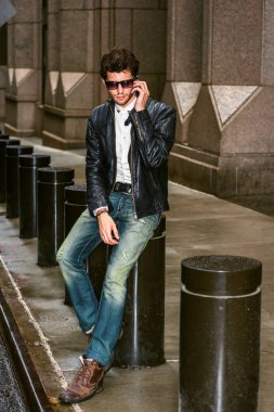 Siyah deri ceketli giyinme, mavi jeans, kahverengi önyükleme ayakkabıların güneş gözlüğü, New York, sokakta oturan sakallı bir genç Avrupa işadamı, cep telefonu üzerinde ara çalışma sırasında konuşurken.