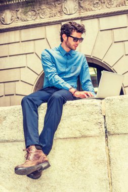 Seyahat, New York'ta çalışma Avrupa iş adamı. Üstünde mavi gömlek, pantolon, çizme ayakkabı, güneş gözlüğü, sakallı, sokakta oturan, okuma, dizüstü bilgisayar üzerinde çalışan genç adam. Filtre uygulanmış etkisi.