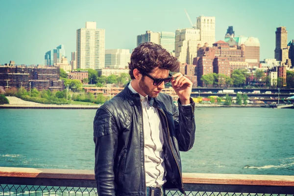 European businessman traveling on East River in New York. Wearing leather jacket, sunglasses, young guy with beard, looking down, sad, thinking, missing family, friends, home. Brooklyn on background.