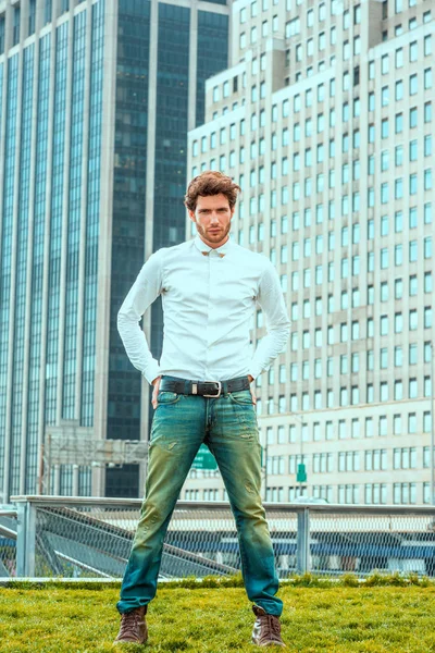 ヨーロッパのビジネスマン旅行 ニューヨークで仕事します 腰に休んで手を身に着けている白いシャツ ブルー ジーンズ 茶色のブート靴 ビジネス地区 緑の芝生の上に立って ひげと若い男 — ストック写真