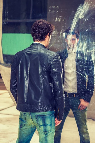 Mysterious Man looking at mirror. A young handsome guy with beard, standing by metal mirror wall, looking at the reflection, thinking. Concept of self assured, self esteem, self checking strategies.