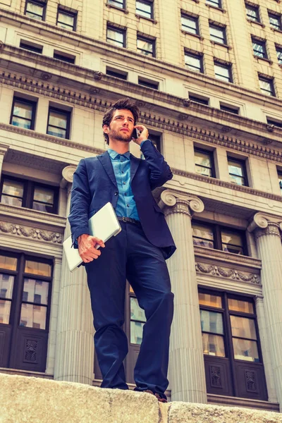 Power of Technology in our modern daily life. Dressing in blue suit, a young European Businessman with beard standing outside in New York, arm carrying laptop computer, talking on mobile phone.