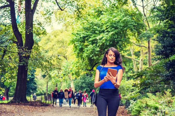 Young American Woman traveling at Central Park, New York, wearing blue sleeveless, off shoulder top, black pants, carrying bag on shoulder, reading tablet computer, walking on road with green trees