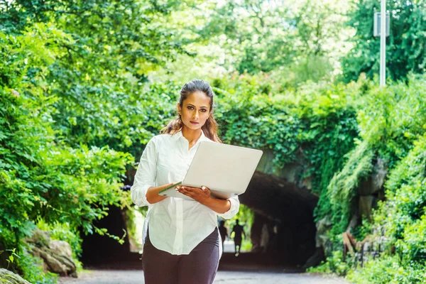 若い東インド アメリカ人女性旅行 ニューヨークで働いている 白いシャツ 黒いズボンを身に着けている保持ラップトップ コンピューター 中央公園でウォーキングします 背景の緑の葉の通り橋 — ストック写真