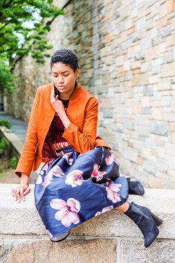 Genç Yalnız Afrikalı-Amerikalı kız New York'ta dışında cep telefonuyla konuşurken, turuncu kırmızı ceketli koyu mor çiçek skeç, siyah çizme ayakkabı, bacaklar, taş çit oturan bükme desenli