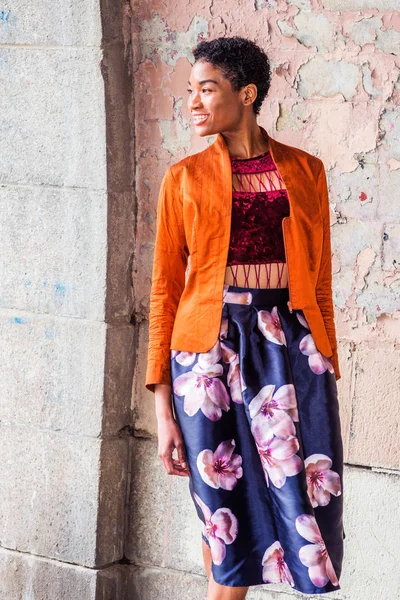 Portrait of Young African American Woman in New York City, with short afro hair, wearing fashionable orange red jacket, dark red under top, dark blue flower patterned skirt, standing by painted wall