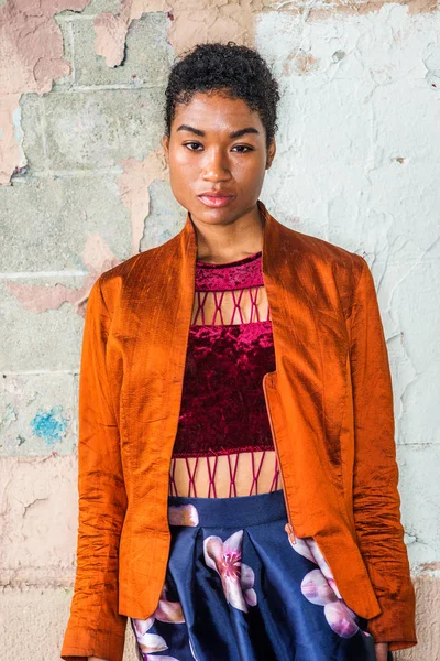 Portrait of Young African American Woman in New York City, with short afro hair, wearing fashionable orange red jacket, dark red under top, dark blue flower patterned skirt, standing by painted wall