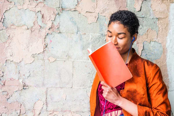 Young African American Woman reading book, listening music outside in New York, wearing orange red jacket, blue earphone, holding red book, standing by painted wall on street, closing eyes, enjoying