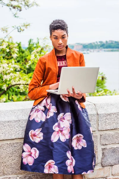 Young African American College Student working on laptop computer outside in New York City, wearing orange red jacket, dark purple flower patterned skirt, standing by stone fence by Hudson River