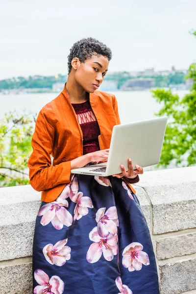 Young African American College Student working on laptop computer outside in New York City, wearing orange red jacket, dark purple flower patterned skirt, standing by stone fence by Hudson River.
