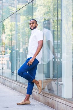 Sakallı, Genç Afrikalı-Amerikalı giyen beyaz kısa kollu gömlek, mavi pantolon, rahat ayakkabı, Gözlük, cam duvar Middletown, Manhattan, New York'taki Caddesi üzerine düşünceler ile karşı duran adam