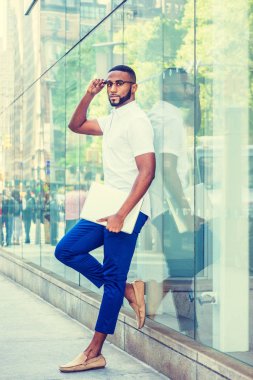Sakallı, genç Afro-Amerikan beyaz kısa kollu gömlek, mavi pantolon, rahat ayakkabı, Gözlük, dizüstü bilgisayar, Manhattan, New York City'de sokakta cam duvara ayakta tutan giyen adam