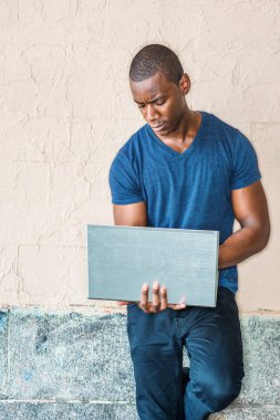 Genç yakışıklı Afrikalı-Amerikalı adam mavi V boyun giyen dizüstü bilgisayarda dışında T gömlek, wall Street karşı ayakta, aşağı bakıyor, okuma, yazma çalışma.