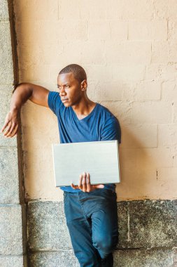 Genç yakışıklı Afrikalı-Amerikalı adam wall Street karşı duran, kol duvara karşı yükselterek, uzağa arıyorsunuz, düşünme T shirt, New York City, mavi V boyun giyen dışında dizüstü bilgisayar üzerinde.