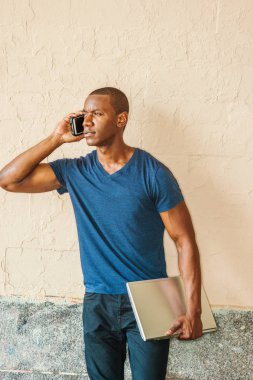 Genç yakışıklı Afrikalı-Amerikalı adam New York'ta mavi V boyun T gömlek giymiş dizüstü bilgisayar, street, duvara karşı ayakta tutan dışında cep telefonuyla konuşurken, dinleme arıyorsunuz