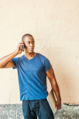 Genç yakışıklı Afrikalı-Amerikalı adam New York'ta mavi V boyun T gömlek giymiş dizüstü bilgisayar, street, duvara karşı ayakta tutan dışında cep telefonuyla konuşurken, dinleme arıyorsunuz