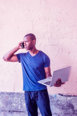 Genç Afro-Amerikan mavi V boyun T gömlek giymiş, wall Street New York'ta tarafından ayakta, dizüstü bilgisayar holding, uzağa arıyorsunuz, cep telefonuyla konuşan adam. Mor Yıllar Proton loo filtre